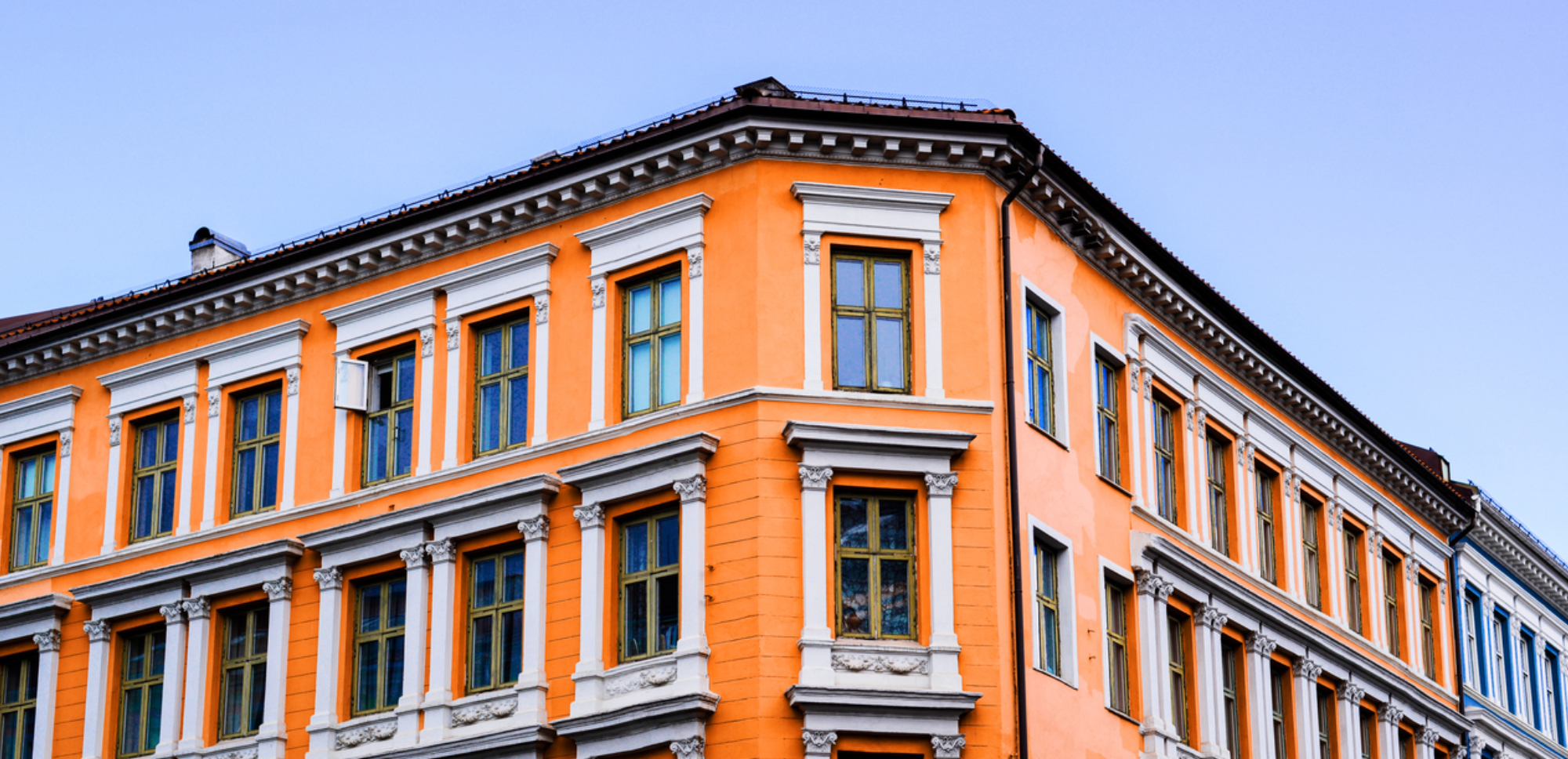 Corner of a classic building with an orange façade, white-framed windows and an ornate cornice against a blue sky.